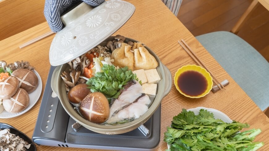 テーブルの上に卓上コンロが置かれ、その上に土鍋が置かれている。土鍋の中には鱈や、豆腐、巾着など、寄せ鍋の具が入っている。