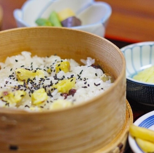 蒸籠に入ったさつまいものおこわ。周りには煮物や漬物などのおかずが小鉢に入っている。